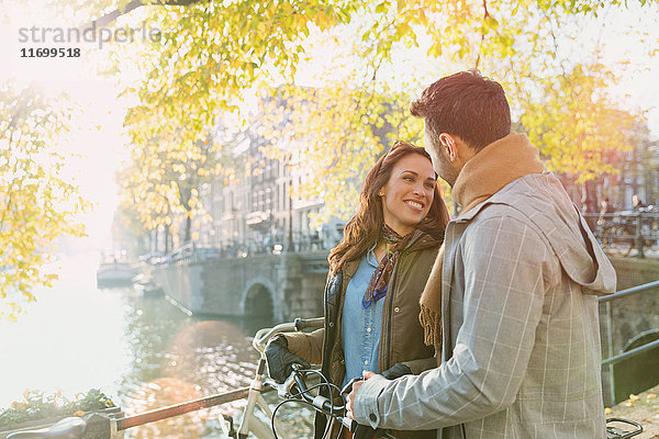 Lächelndes junges Paar mit Fahrrad auf der Brücke über den sonnigen Herbstkanal in Amsterdam
