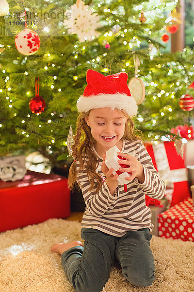 Lächelndes Mädchen mit Weihnachtsmannmütze öffnet ein Geschenk vor dem Weihnachtsbaum