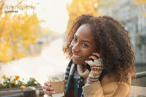 Lächelnde junge Frau trinkt Kaffee und telefoniert im Herbst im Straßencafé.