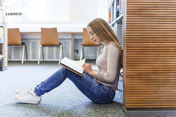 Teenagermädchen sitzt auf dem Boden in einer öffentlichen Bibliothek und liest Bücher.