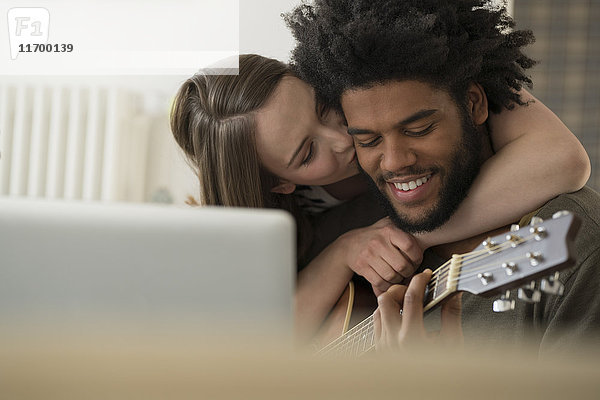 Nahaufnahme einer Frau  die einen lächelnden Mann küsst  der vor dem Laptop Gitarre spielt.