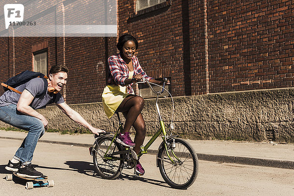Junge Frau auf dem Fahrrad zieht jungen Mann  stehend auf Skateboard