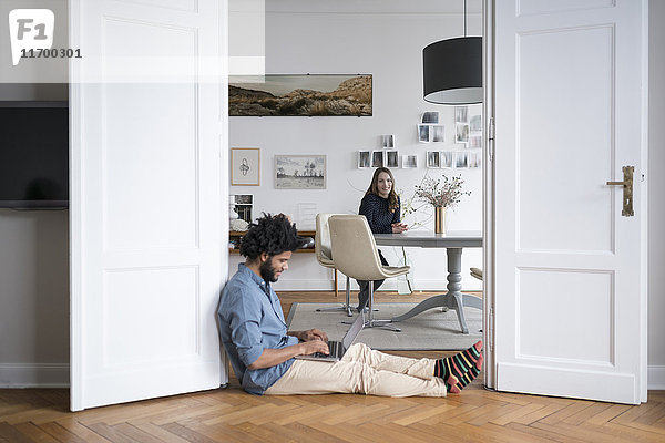 Mann zu Hause am Boden sitzend mit Laptop und Frau im Hintergrund