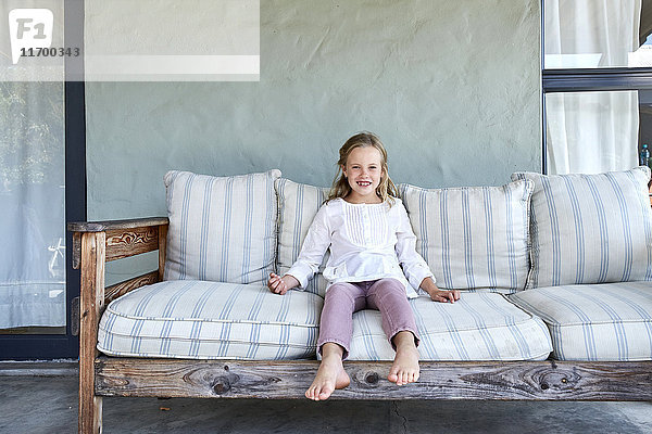 Porträt eines lächelnden blonden Mädchens auf der Couch auf der Terrasse