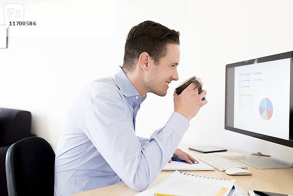 Lächelnder Geschäftsmann mit einer Tasse Kaffee  der das Kuchendiagramm auf dem Computerbildschirm analysiert.