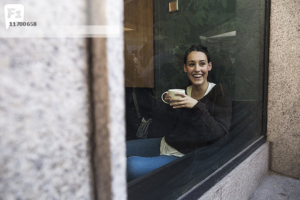 Porträt einer lächelnden Frau mit einer Tasse Kaffee hinter einer Fensterscheibe in einem Coffee-Shop.