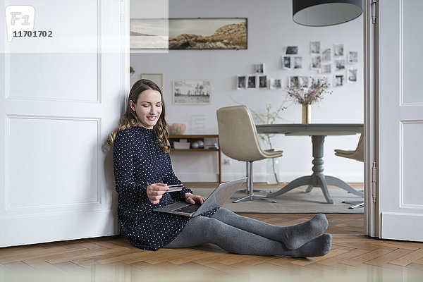 Lächelnde Frau zu Hause auf dem Boden sitzend mit Laptop im Türrahmen mit Kreditkarte