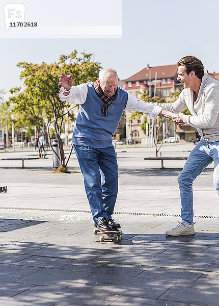 Erwachsener Enkel  der dem älteren Mann auf dem Skateboard assistiert.