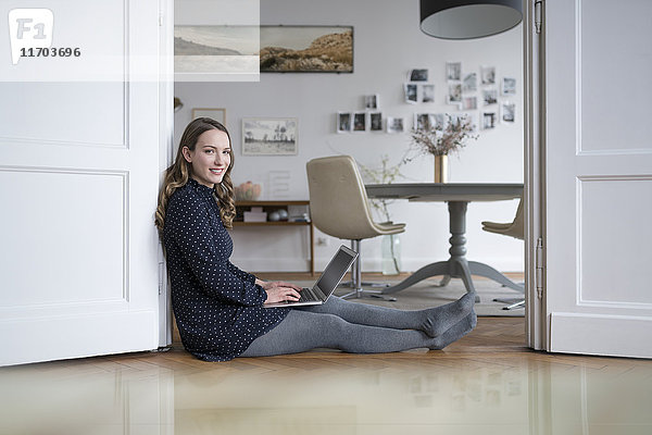 Lächelnde Frau zu Hause auf dem Boden sitzend  arbeitet mit Laptop im Türrahmen