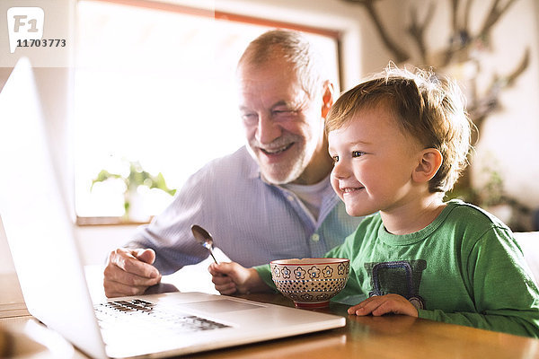 Großvater und Enkel sitzen am Tisch  essen und benutzen Laptop
