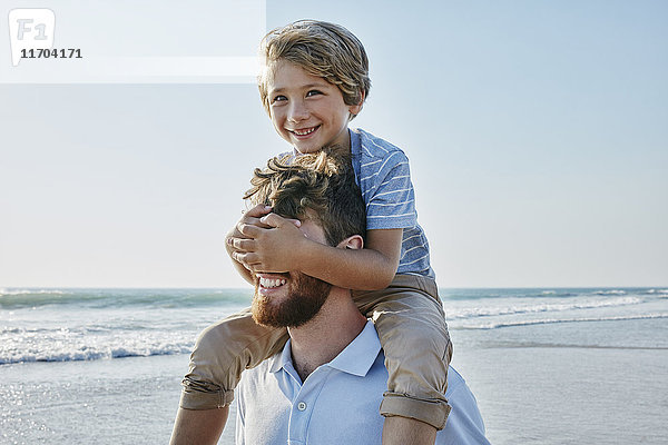 Vater trägt Sohn Huckepack am Strand  Sohn bedeckt Vater's Augen
