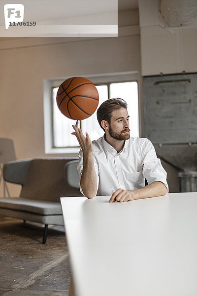 Porträt eines nachdenklichen jungen Geschäftsmannes  der am Tisch in einem Loft sitzt und Basketball auf seinem Finger balanciert.