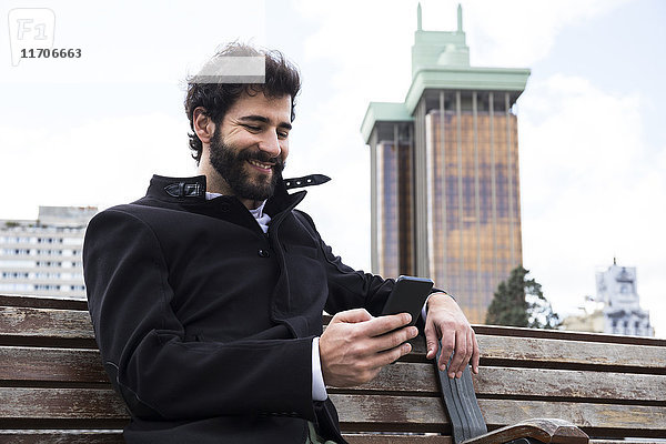 Spanien  Madrid  lächelnder junger Mann mit Vollbart auf einer Bank sitzend mit Blick auf das Handy