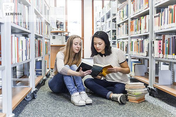 Zwei Teenager-Mädchen sitzen auf dem Boden in einer öffentlichen Bibliothek und lesen in einem Buch.
