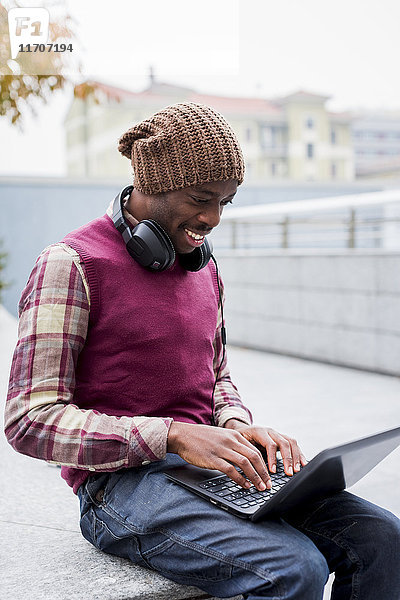 Lächelnder Mann mit Kopfhörer auf der Bank sitzend mit Laptop