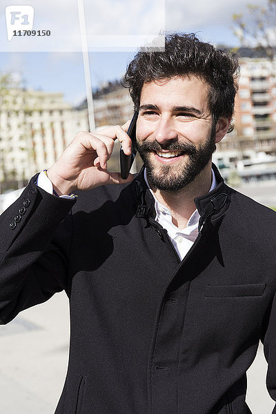 Porträt eines lächelnden jungen Mannes mit Vollbart am Telefon
