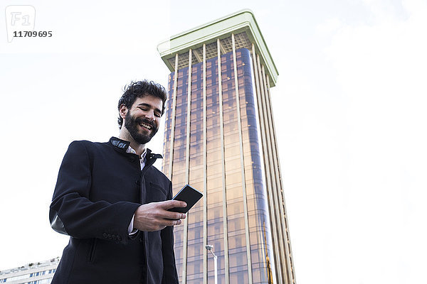 Spanien  Madrid  lächelnder junger Mann mit Blick auf sein Smartphone vor dem Wolkenkratzer