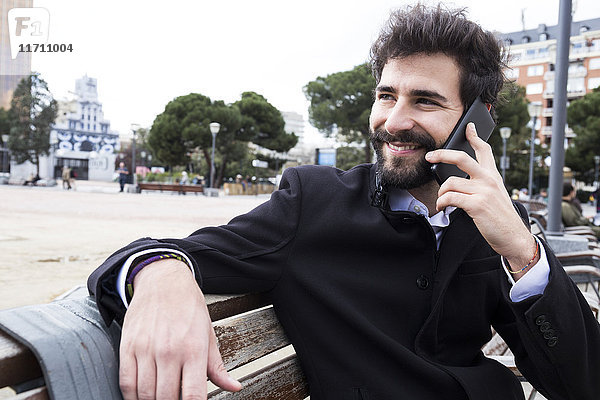 Porträt eines lächelnden jungen Mannes mit Vollbart am Telefon  der auf einer Bank sitzt.