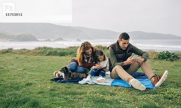 Familie mit Hund  der an der Küste auf einer Decke sitzt und drahtlose Geräte benutzt.