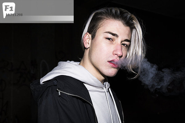 Porträt eines jungen Mannes beim Ausatmen von Zigarettenrauch vor schwarzem Hintergrund
