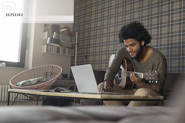 Mann sitzt im Wohnzimmer auf dem Sofa und spielt Gitarre vor dem Laptop.