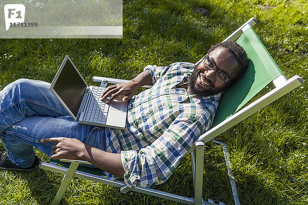 Lächelnder Mann mit Laptop auf Liegestuhl auf einer Wiese mit Blick zur Kamera
