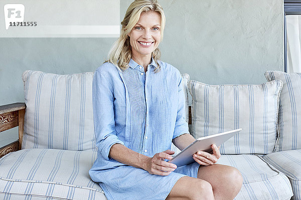 Porträt einer lächelnden blonden Frau auf der Couch mit Tablette