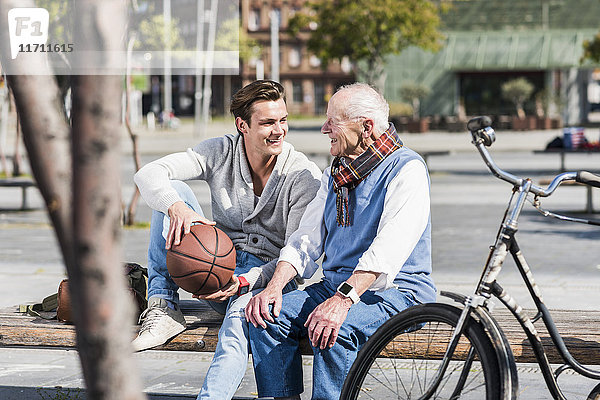 Senior Mann und erwachsener Enkel mit Basketball auf einer Bank im Gespräch