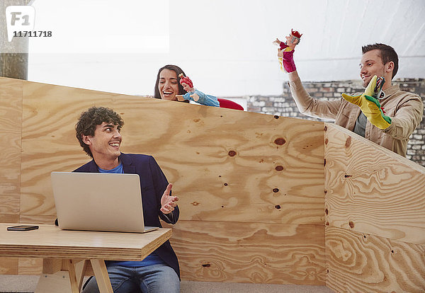 Junger Geschäftsmann arbeitet am Laptop mit Kollegen  die hinter einer Holzwand Handpuppen spielen.