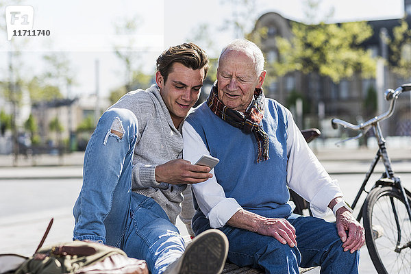 Senior Mann und erwachsener Enkel auf einer Bank mit Blick auf Handy