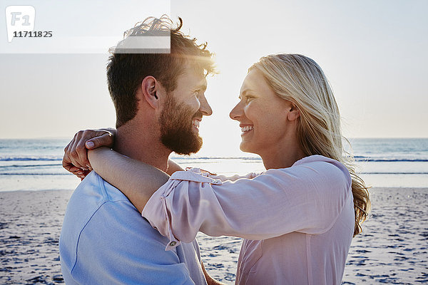 Lächelndes Paar  das sich bei Sonnenuntergang am Strand umarmt.