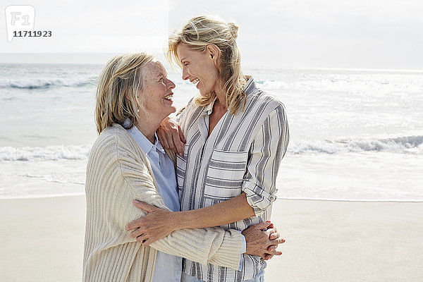 Seniorin und ihre erwachsene Tochter stehen am Strand und umarmen sich.