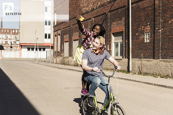 Junges Paar  das auf der Straße Fahrrad fährt  Frau  die auf einem Gestell steht und sich selbst mitnimmt.
