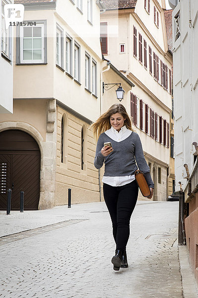 Deutschland  Tübingen  lächelnder junger Student beim Gehen auf das Handy schauen