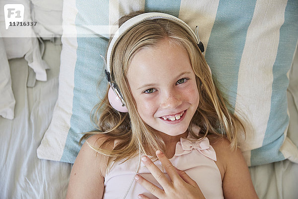Porträt eines lächelnden kleinen Mädchens  das zu Hause auf dem Bett liegt und Musik mit Kopfhörern hört.