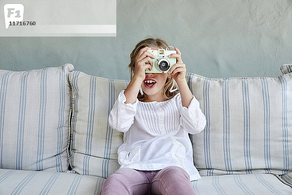 Porträt eines lächelnden kleinen Mädchens  das auf der Couch sitzt und mit der Kamera fotografiert.
