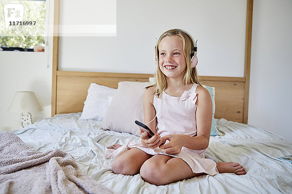 Porträt eines lächelnden kleinen Mädchens  das auf dem Bett sitzt und mit Kopfhörern Musik hört.