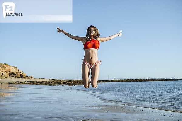 Lächelnde junge Frau  die am Strand in die Luft springt.