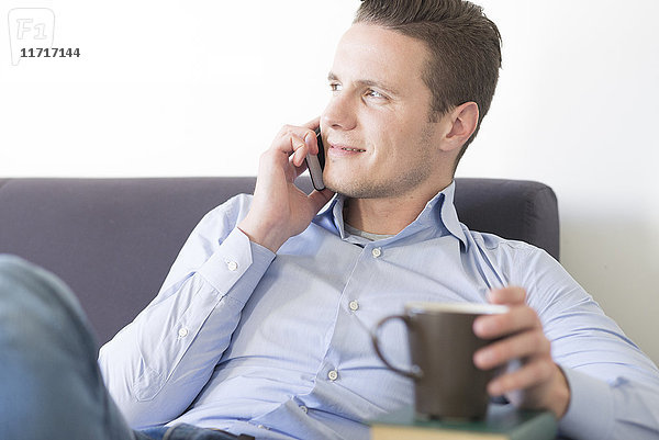 Lächelnder Geschäftsmann auf der Couch mit Smartphone und Tasse Kaffee