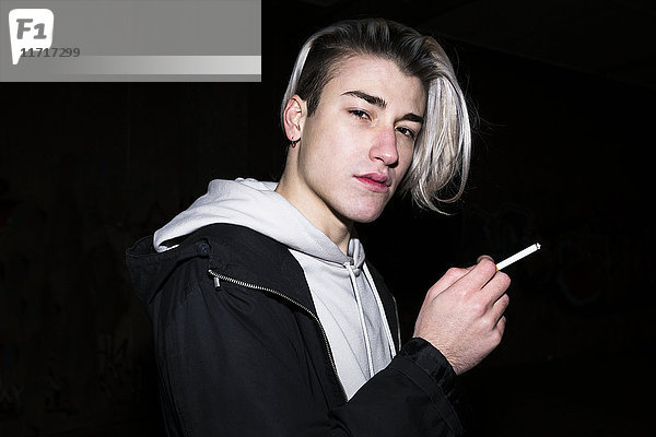 Porträt eines jungen Mannes  der vor schwarzem Hintergrund eine Zigarette raucht.