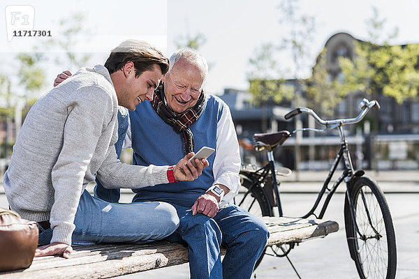 Senior Mann und erwachsener Enkel auf einer Bank mit Blick auf Handy
