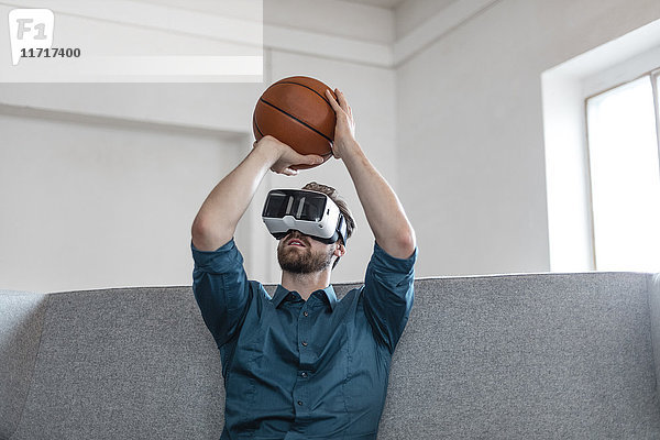 Junger Mann mit Basketball auf der Couch sitzend mit Virtual Reality Brille