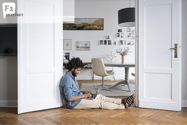 Mann zu Hause auf dem Boden sitzend  arbeitet mit Laptop im Türrahmen