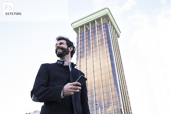 Spanien  Madrid  lächelnder junger Mann mit Smartphone vor Wolkenkratzer