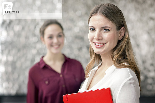 Portrait der lächelnden jungen Geschäftsfrau mit Notizbuch und Kollegin im Hintergrund