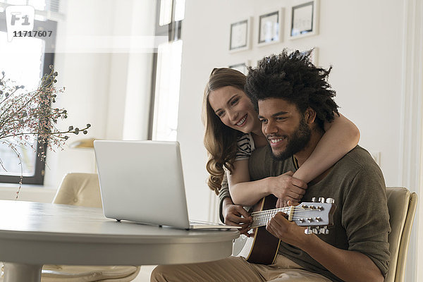 Lächelndes Paar vor dem Laptop im Wohnzimmer  Mann beim Gitarrespielen