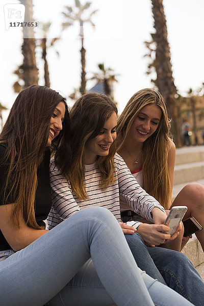 Drei lächelnde junge Frauen sitzen auf der Treppe und schauen aufs Handy.