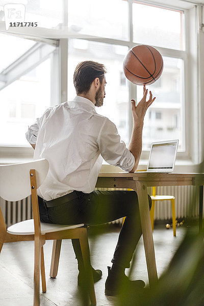 Rückansicht eines jungen Mannes am Schreibtisch mit Blick auf den Laptop beim Balancieren eines Basketballs am Finger