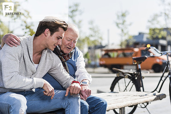 Senior Mann und erwachsener Enkel auf einer Bank mit Blick auf ihre Smartuhren