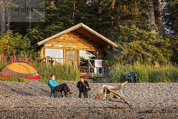 Zwei Frauen entspannen sich in Stühlen am Ufer von Hesketh Island mit einer öffentlich zugänglichen Hütte im Hintergrund  Southcentral Alaska  USA